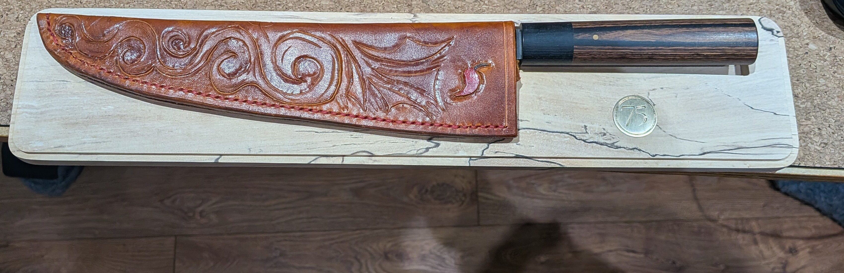 The knife in its leather sheath on the spalted maple bed, before finishing – the brass disc engraved with 75 is visible next to the handle